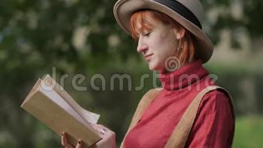 年轻迷人的红头发女人戴着帽子和毛衣在一个自然公园看书。 秋天凉爽的一天。